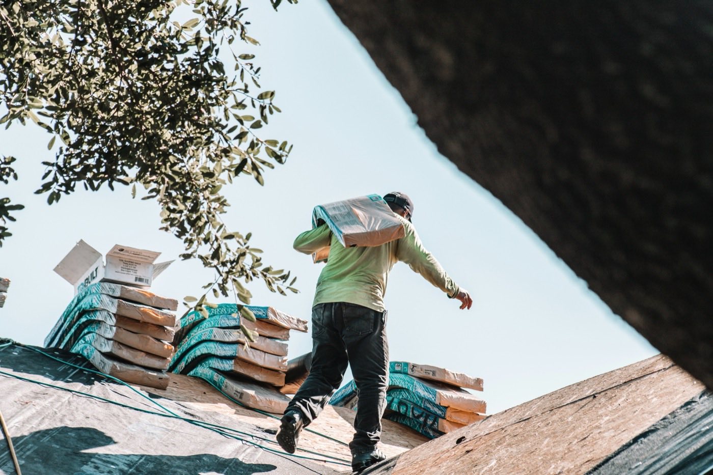 A roofer carrying a bundle of shingles up a roof under a clear sky